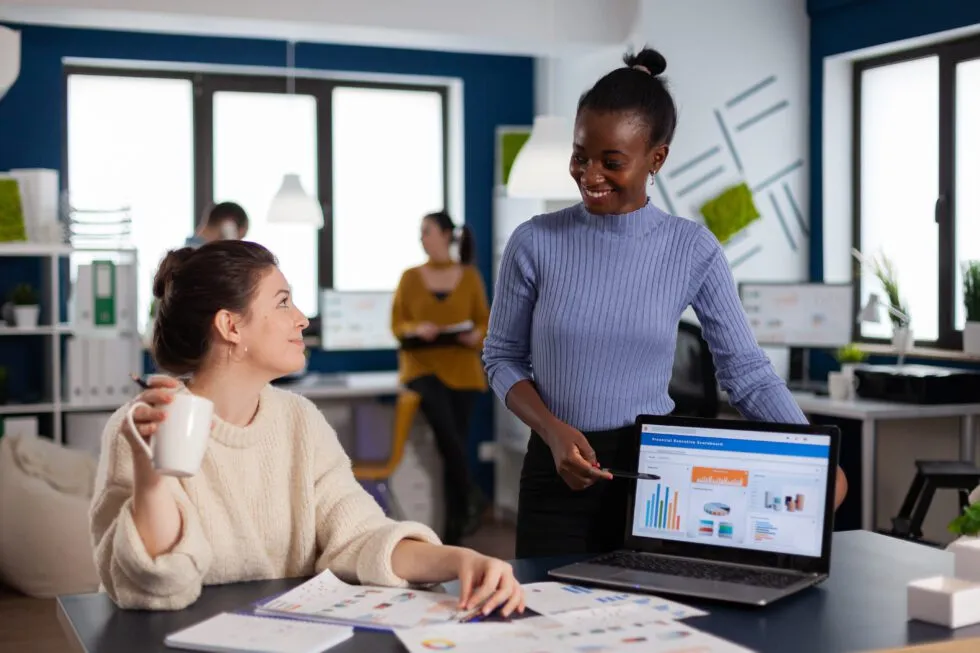 4-3-980×653 A group of professionals collaborates in a modern office, discussing data and charts displayed on a laptop at a table.